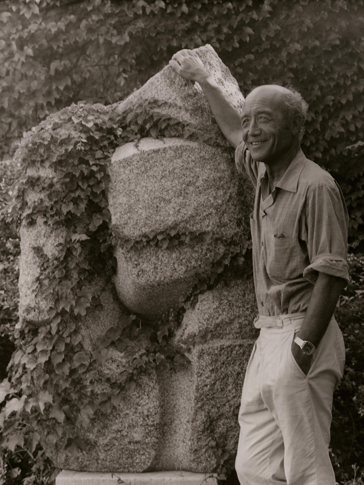 Isamu Noguchi sorrindo em seu jardim no Museu Noguchi, Long Island City, com a mão em um grande...